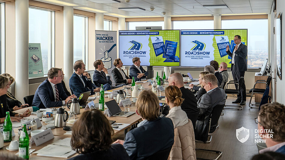 Raumansicht mit länglichem Tisch, an denen Menschen vor einer Präsentation sitzen