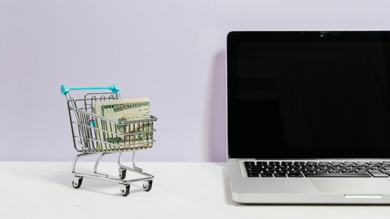 eCommerce sicher gestalten Miniatureinkaufswagen in dem ein Geldschein ist neben einem Laptop