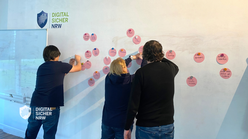 DIGITAL.SICHER.NRW Workshop – Personen stehen vor einer Whiteboard-Wand und kleben Notizen ans Board