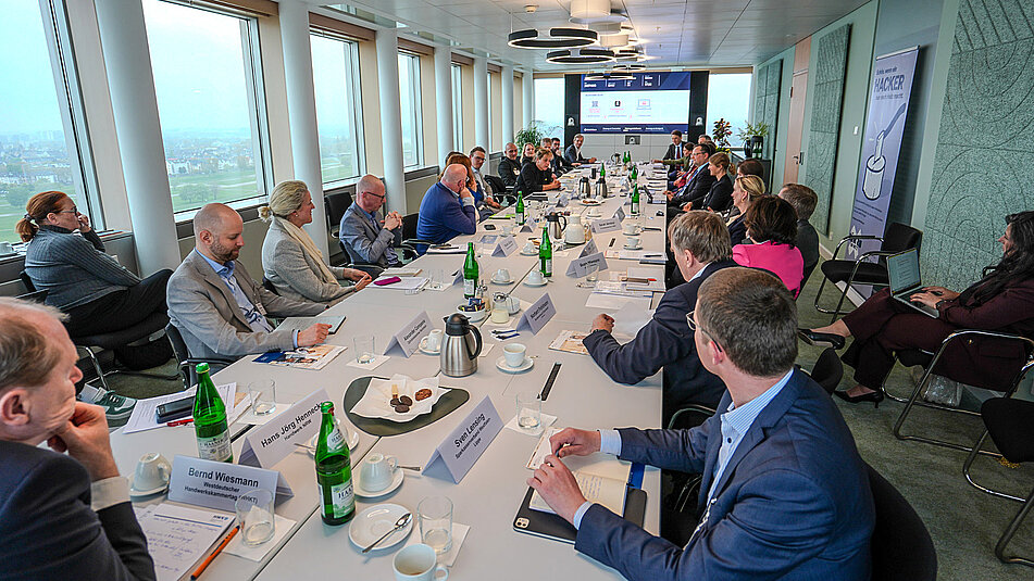 Foto der Teilnehmenden an langem Tisch mit Blick auf den Präsentationsbildschirm